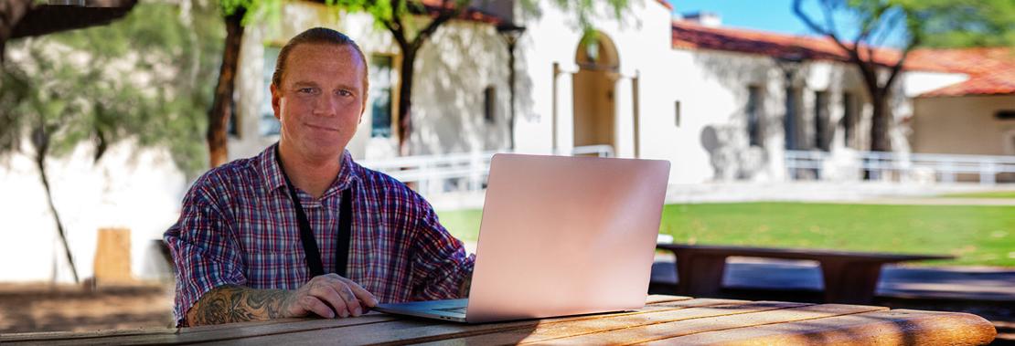 彼得·基思坐在市中心校园外的一张桌子旁，手里拿着一台笔记本电脑